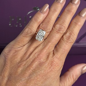 Pu&ograve; includere: Un anello color argento con una grande pietra rettangolare trasparente sfaccettata, montata su una struttura a griffe. L'anello &egrave; indossato su un dito, mettendo in risalto la gemma prominente che riflette la luce con un aspetto brillante.