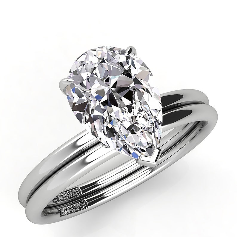 Puede incluir: Un anillo de compromiso de diamante de forma pera con una banda de boda a juego. Ambas bandas son de oro blanco y llevan la inscripci&oacute;n "SABEHI" en el interior.