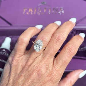 Puede incluir: Un anillo de compromiso de oro rosa con una gran piedra central de diamante de talla ovalada y un halo de diamantes m&aacute;s peque&ntilde;os. El anillo est&aacute; en una mano con esmalte de u&ntilde;as blanco. El fondo incluye una caja morada.