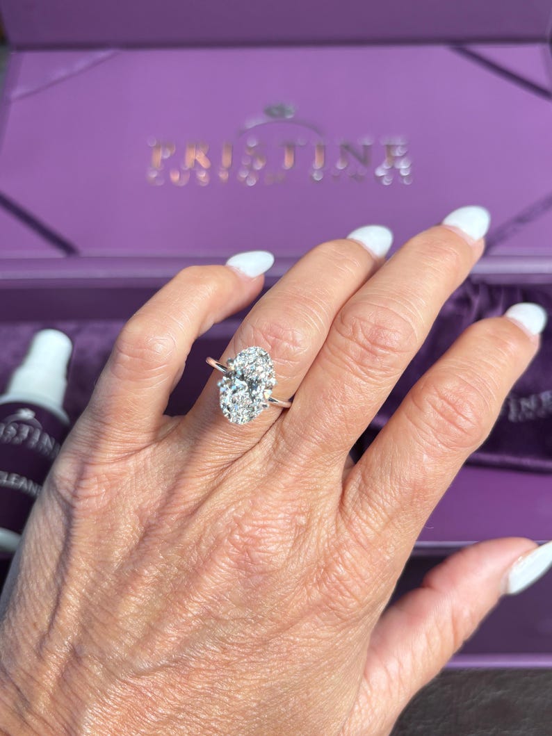Puede incluir: Un anillo de compromiso de oro rosa con una gran piedra central de diamante de talla ovalada y un halo de diamantes m&aacute;s peque&ntilde;os. El anillo est&aacute; en una mano con esmalte de u&ntilde;as blanco. El fondo incluye una caja morada.
