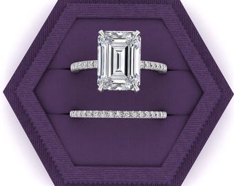 May include: A diamond engagement ring with an emerald cut center stone and a diamond band, along with a matching diamond wedding band, presented in a purple velvet hexagonal ring box.