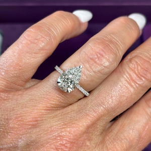 Può includere: Un anello di fidanzamento con diamante a forma di pera e fascia pavé. L'anello presenta un grande diamante centrale trasparente e diamanti più piccoli incastonati lungo la fascia. L'anello è in metallo bianco, probabilmente platino o oro bianco.
