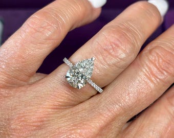 Puede incluir: Un anillo de compromiso de diamantes con una piedra central en forma de pera y una banda pavimentada. El anillo presenta un diamante grande y claro y diamantes más pequeños engastados en la banda. El anillo está engastado en metal blanco, probablemente platino u oro blanco.