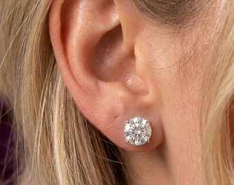May include: A close-up of a woman's ear with a large, round, sparkling diamond stud earring. The earring is set in a simple, classic design. The woman has blonde hair.