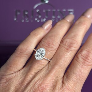 K&ouml;nnte beinhalten: Ein silberfarbener Ring mit einem gro&szlig;en, l&auml;nglichen Stein im Marquise-Schliff, der von einem Kranz aus kleineren, rund geschliffenen Steinen umgeben ist. Der Ring sitzt auf einer schlichten, d&uuml;nnen Metallschiene. Im Hintergrund steht PRISTINE.