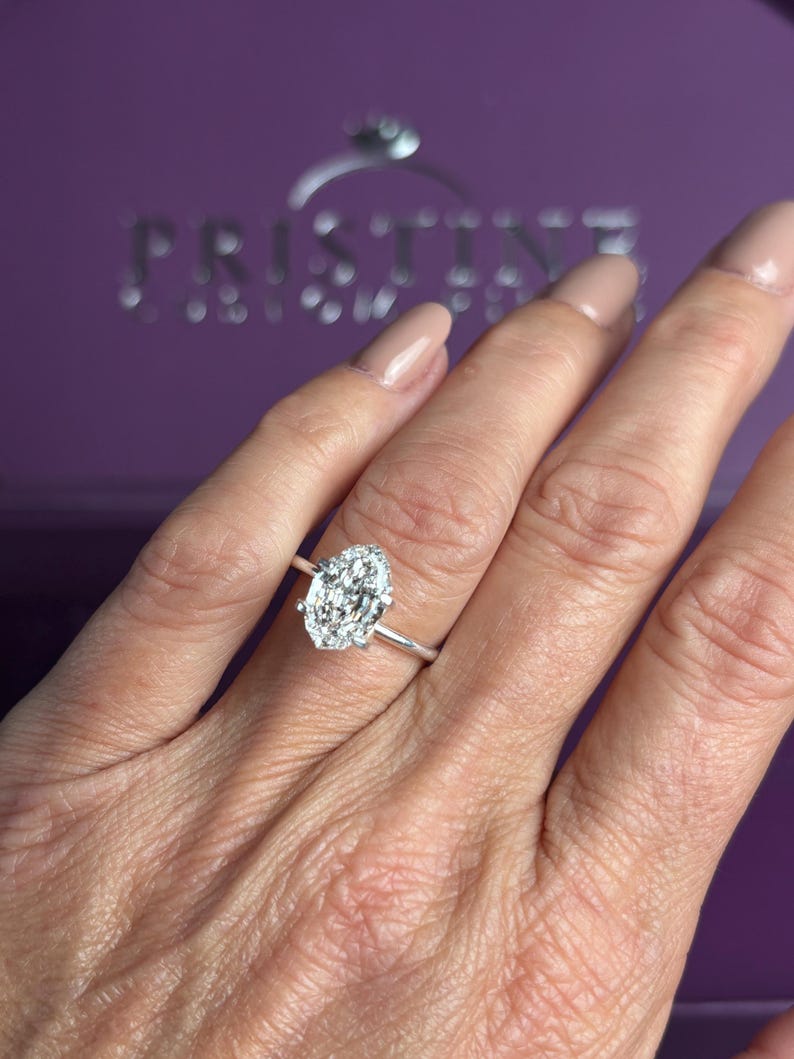 K&ouml;nnte beinhalten: Ein silberfarbener Ring mit einem gro&szlig;en, l&auml;nglichen Stein im Marquise-Schliff, der von einem Kranz aus kleineren, rund geschliffenen Steinen umgeben ist. Der Ring sitzt auf einer schlichten, d&uuml;nnen Metallschiene. Im Hintergrund steht PRISTINE.