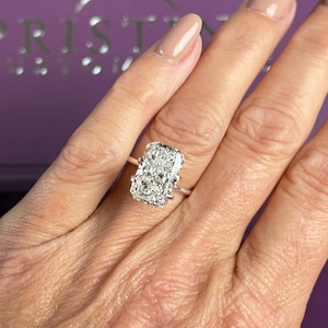 Pu&ograve; includere: Un anello con diamante taglio radiante caratterizzato da una grande pietra rettangolare trasparente a faccette multiple, montata su una semplice fascia in metallo color argento lucido con quattro griffe che fissano la pietra centrale.