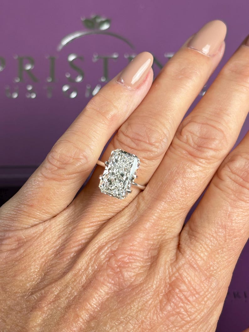 Pu&ograve; includere: Un anello con diamante taglio radiante caratterizzato da una grande pietra rettangolare trasparente a faccette multiple, montata su una semplice fascia in metallo color argento lucido con quattro griffe che fissano la pietra centrale.
