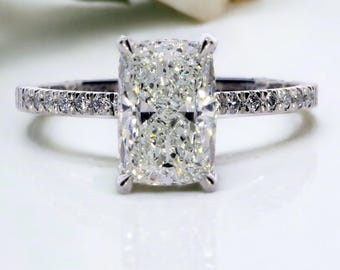 May include: A radiant-cut diamond engagement ring. The large, rectangular-shaped diamond is the focal point, set in a platinum band. The band is embellished with smaller, pave-set diamonds, adding sparkle. The ring is displayed on a white surface.