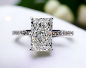 May include: A silver engagement ring with a large, clear, rectangular-shaped diamond. The diamond is set in a prong setting and the band is adorned with smaller diamonds.
