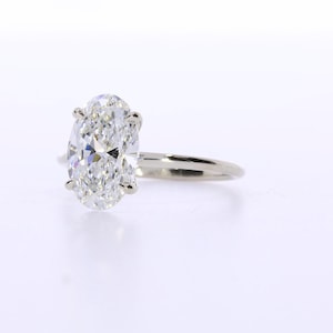 May include: A solitaire engagement ring featuring an oval-cut diamond. The diamond is set in a four-prong setting on a thin, polished, white gold band. The ring is displayed against a white background.