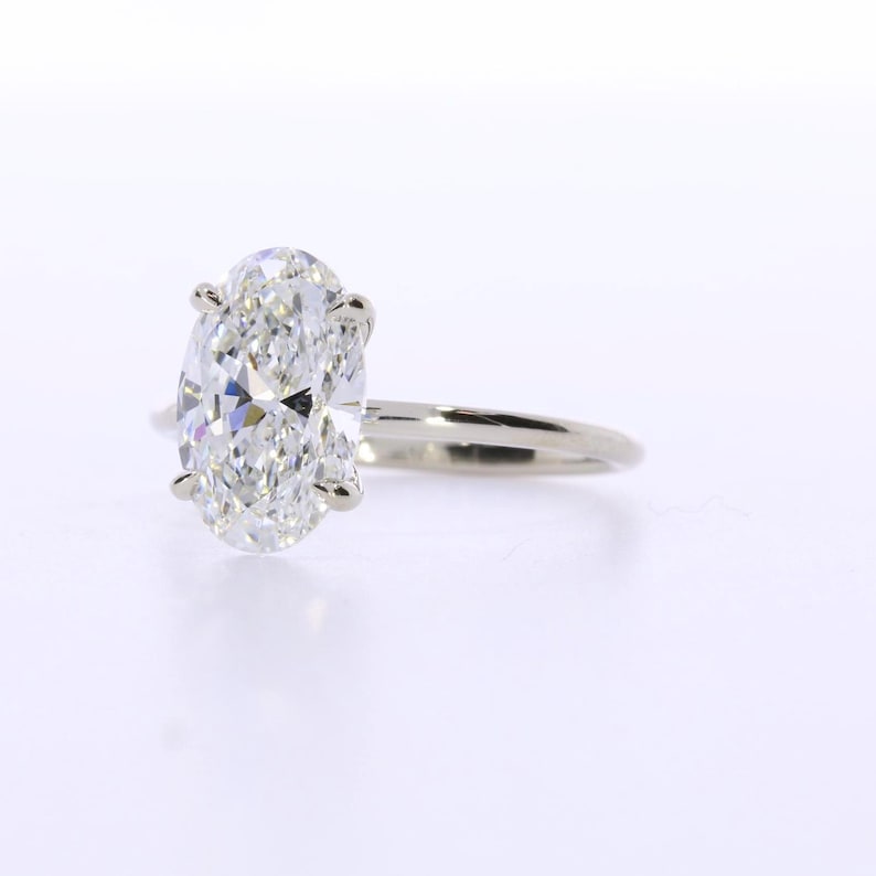 May include: A solitaire engagement ring featuring an oval-cut diamond. The diamond is set in a four-prong setting on a thin, polished, white gold band. The ring is displayed against a white background.
