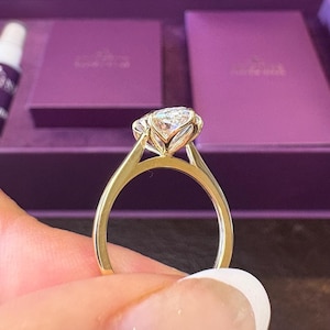 May include: A gold ring featuring a large, clear, faceted round gemstone held in a six-prong setting. The band is smooth and polished, showcasing a classic design with a high-profile setting that elevates the stone above the metal band.