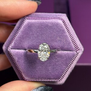 May include: A diamond engagement ring with an oval-cut center stone. The ring is set in a silver band and presented in a purple velvet ring box. The ring box is hexagonal in shape.