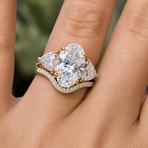 May include: A gold engagement ring set featuring a large oval-cut center stone flanked by two trillion-cut side stones, paired with a curved wedding band encrusted with small round pavé diamonds along the entire top edge of the band.