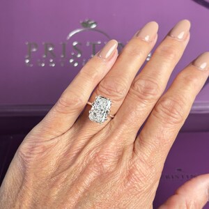 Pu&ograve; includere: Un anello di fidanzamento con diamante taglio radiante, caratterizzato da una grande pietra centrale rettangolare con molteplici sfaccettature montata su una sottile fascia in metallo lucido. Il testo PRISTINE &egrave; visibile sullo sfondo viola.