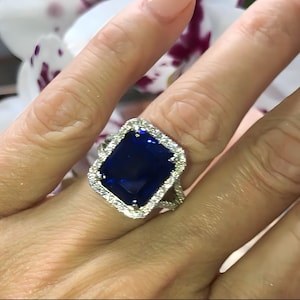Puede incluir: Un anillo de plata con una gran piedra preciosa rectangular de color azul oscuro rodeada por un halo de piedras preciosas más pequeñas y transparentes.