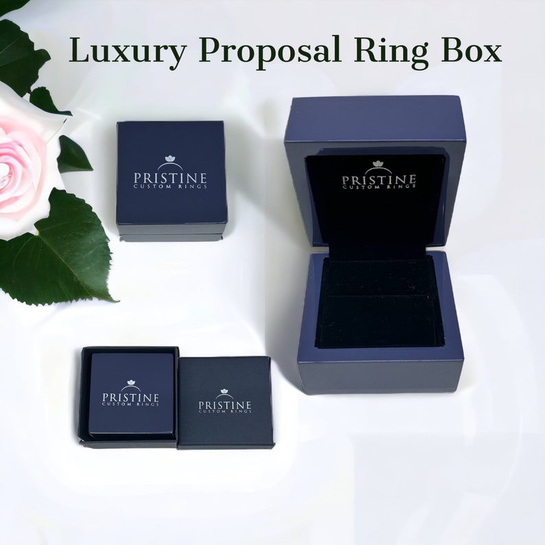 May include: A dark blue ring box with a velvet interior. The box is open and the lid has the text "PRISTINE CUSTOM RINGS" printed on it.