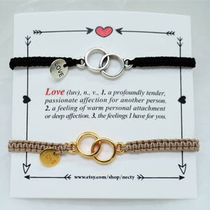 Op de afbeelding: Twee vriendschapsarmbanden met een zilveren en gouden, in elkaar grijpende cirkelbedel. De armbanden zijn van zwart en bruin koord met een kleine ronde bedel waarop "LOVE" staat. De armbanden worden gepresenteerd op een witte kaart met de definitie van het woord "love" erop gedrukt.