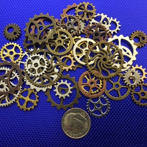 May include: A collection of various sized metal gears in bronze, gold, and silver tones, scattered on a textured blue surface. A dime is included for size reference. The gears have intricate designs and are suitable for steampunk or craft projects.