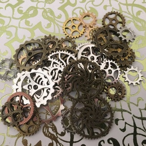 May include: A collection of assorted metal gears and cogs in various sizes and finishes, including antique bronze, silver, copper, and gold tones. The pieces feature intricate cut-out designs, spokes, and teeth, scattered in a cluster.