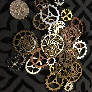 May include: A collection of assorted metal gear and cog charms in various sizes and metallic finishes including gold, silver, copper, and bronze, displayed alongside a United States one dime coin for scale, featuring intricate cut-out designs.