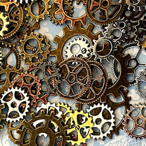 May include: A collection of assorted metal gears and cogs in various sizes and finishes, including antique bronze, copper, and silver tones. The pieces feature intricate teeth and spoke designs, layered together to create a dense, mechanical pattern.