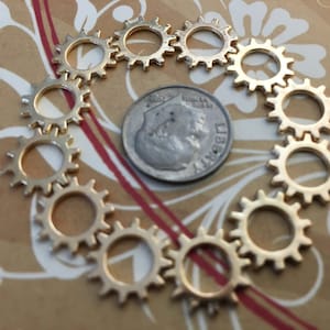 May include: A collection of twelve small, circular, gold-toned metal gear charms arranged in a ring formation around a single United States dime, which serves as a scale reference for the size of the individual gear components.