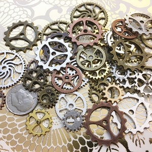 May include: A collection of various metal gears in bronze, copper, silver, and gold tones. The gears vary in size and design, with some having intricate cut-out patterns. A dime is included for size reference. The gears are arranged on a gold patterned surface.
