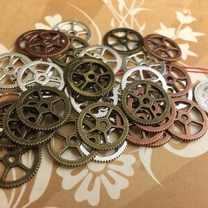 May include: A collection of small, circular metal gear charms in assorted finishes including antique bronze, copper, and silver. The gears feature various spoke designs and teeth along the outer edges. A United States dime is included for scale.