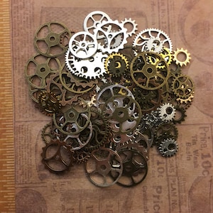 May include: A collection of small metal gears in various sizes and colors, including silver, gold, and bronze. The gears are scattered on a brown background with printed text and a ruler, suitable for steampunk or craft projects.