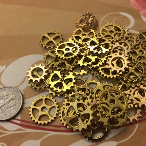 May include: A collection of small, gold-colored metal gear-shaped charms with various intricate cut-out designs. A silver-colored United States dime coin is positioned to the left of the pile to provide a visual scale for the size of the gears.