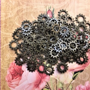 May include: A collection of small, circular bronze-toned metal gear charms with serrated edges. A pink measuring tape is visible on the left side, showing the gears measure approximately 0.5 inches in diameter against a floral background.