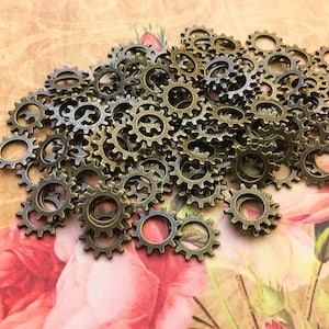 May include: A collection of small, circular, bronze-colored metal gear charms with serrated edges. Each gear features a central hole and distinct teeth around the perimeter, arranged in a loose pile to display their uniform shape and metallic finish.