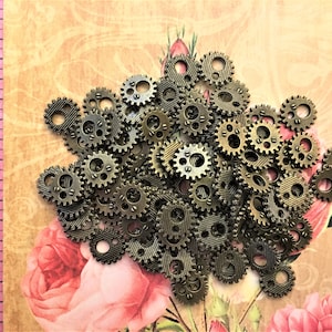 May include: A collection of small, round, bronze-colored metal gear charms with textured surfaces and two circular holes. A pink measuring tape is visible on the left side, showing measurements from 14 to 17 inches for scale reference.