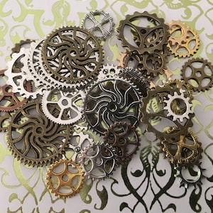 May include: A collection of assorted metal gears and cogs in various sizes and finishes, including antique bronze, silver, copper, and gold tones. The pieces feature intricate cut-out designs, varying tooth counts, and circular shapes.