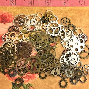 May include: A collection of various-sized metal gears in shades of silver, bronze, and gold. The gears are scattered on a floral patterned background with a pink ruler at the top. The gears are ideal for steampunk or industrial-themed crafts.