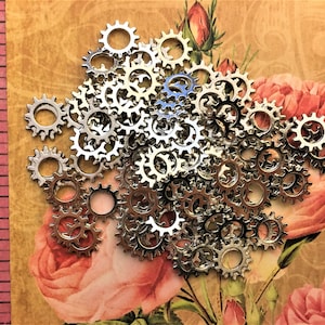May include: A collection of small, silver-toned metal gear charms with serrated edges. A pink measuring tape is visible on the left side, showing the gears measure approximately 0.5 inches in diameter. The charms are arranged in a dense pile.