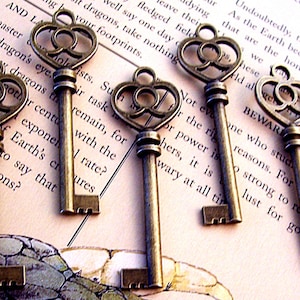 May include: Five antique-style skeleton keys with ornate heart-shaped bows. The keys are made of brass and have a worn, aged finish. The keys are arranged on a page of a book with text.