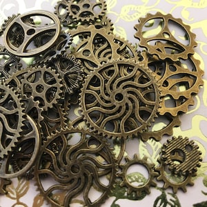 May include: A collection of antique gold-toned metal gears of various sizes and designs. The gears are arranged in a random pattern on a white and gold background.