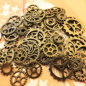 May include: A collection of antique gold-toned metal gears of various sizes and shapes. The gears are arranged in a random pattern on a light brown surface.