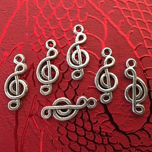 以下が含まれることがあります： 銀色のト音記号チャーム6個のクローズアップ。チャームは、質感のある赤い表面に配置されています。各チャームの上部には、ジュエリーやその他の工芸品に取り付けるためのループが付いています。音楽をテーマにしたチャームです。