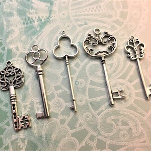 May include: Five antique-style silver keys with ornate designs. The keys vary in shape, including a clover, a heart, and a fleur-de-lis. They are arranged on a patterned background with a ruler along the left side.