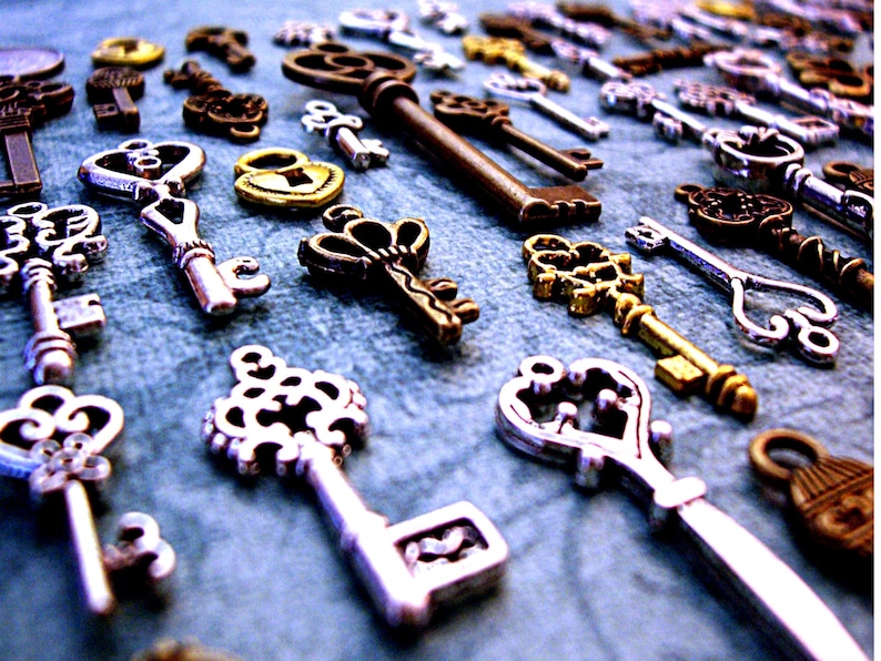 May include: A collection of antique-style keys in silver, gold, and bronze finishes. The keys feature intricate designs and are arranged on a blue surface.