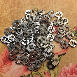 May include: A collection of small, silver-toned metal gear-shaped buttons with a textured, striped surface and two holes for sewing. The buttons are arranged in a pile next to a pink measuring tape showing markings in inches.
