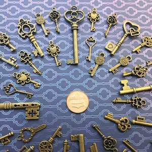 May include: A collection of various antique-style bronze-toned metal key charms arranged in a circular pattern around a single copper-colored United States one-cent coin placed in the center, showcasing diverse ornate bow and bit designs.