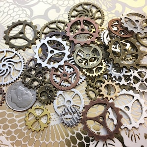 May include: A collection of various metal gears in bronze, copper, silver, and gold tones. The gears are of different sizes and designs, some with intricate patterns. A dime is included for size reference, set against a gold floral background.