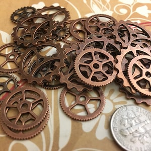 May include: A collection of small, bronze-colored metal gears of varying sizes. The gears have intricate teeth and circular designs, suggesting a steampunk or industrial theme. A US dime is included for size reference.