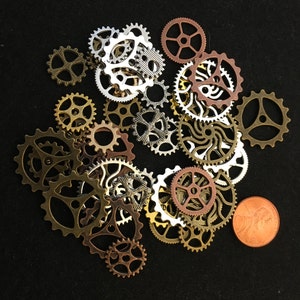 May include: A collection of small metal gears in various colors, including gold, silver, and copper, scattered on a black surface. A copper penny is included for size comparison. The gears have different designs and sizes, suitable for steampunk or craft projects.