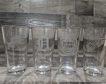 Könnte beinhalten: Vier klare Pintgläser mit geätzten Logos von Detroiter Sportteams: Lions, Tigers, Pistons und Red Wings. Die Gläser sind zylindrisch und stehen auf einer Holzoberfläche.